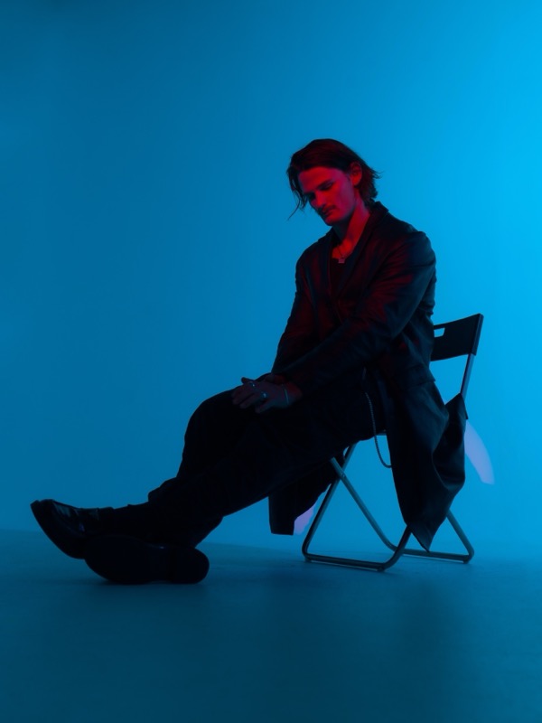 Audent seated on a folding chair in blue studio light, press photo